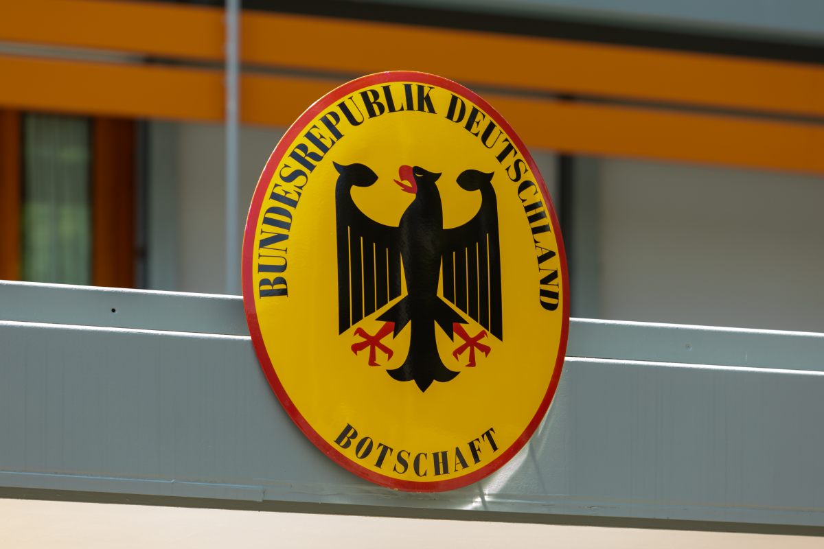 Das Bild zeigt das offizielle Emblem der deutschen Botschaft - ein ovales gelbes Schild mit rotem Rand. In der Mitte befindet sich der deutsche Bundesadler in schwarzer Farbe mit roten Krallen und rotem Schnabel. Um den Adler herum steht in schwarzen Großbuchstaben