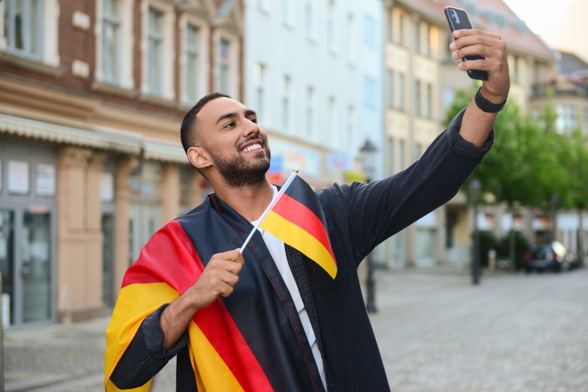 Das Bild zeigt einen jungen Mann mit Bart in dunkler Kleidung, der fröhlich lächelnd ein Selfie mit seinem Smartphone macht. Er hält eine kleine deutsche Flagge in der Hand und trägt die deutsche Fahne (schwarz-rot-gold) um die Schultern. Im Hintergrund ist eine europäische Altstadt mit historischen Gebäuden, gepflasterten Straßen und grünen Bäumen zu sehen. Die Szene vermittelt eine festliche, patriotische Stimmung, möglicherweise während eines Feiertags oder Sportereignisses.