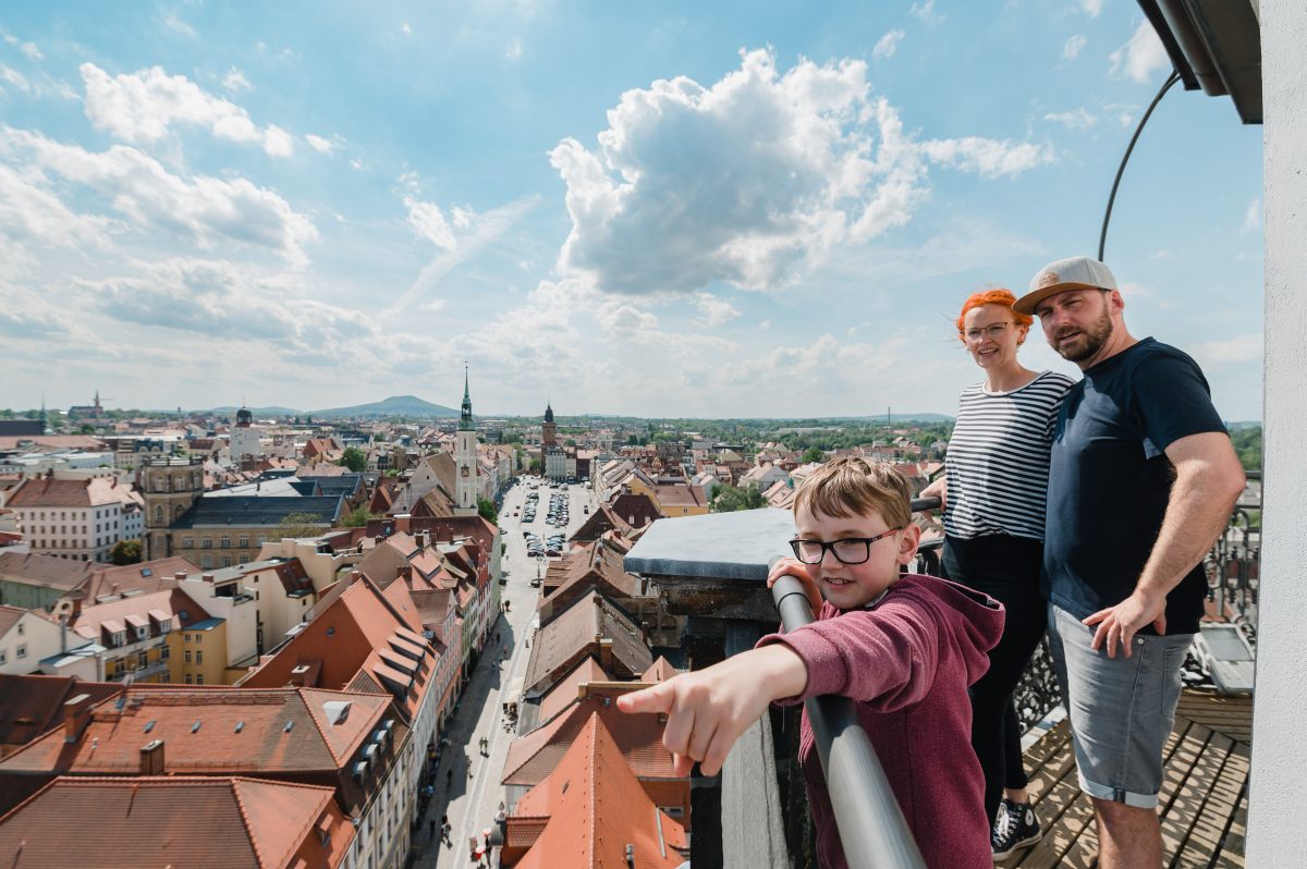 Das Bild zeigt eine Familie mit drei Personen auf dem Görlitzer Rathausturm. Im Vordergrund steht ein Junge mit Brille und bordeauxrotem Pullover, der auf etwas in der Stadt unten zeigt. Dahinter befinden sich ein Mann mit Baseballkappe und dunklem T-Shirt sowie eine Frau mit kurzen roten Haaren in einem gestreiften Oberteil. Sie genießen den Panoramablick über die historische Altstadt mit roten Ziegeldächern, Kirchtürmen und klassischer Architektur. Der Himmel ist teilweise bewölkt und das Wetter wirkt angenehm für einen Familienausflug.