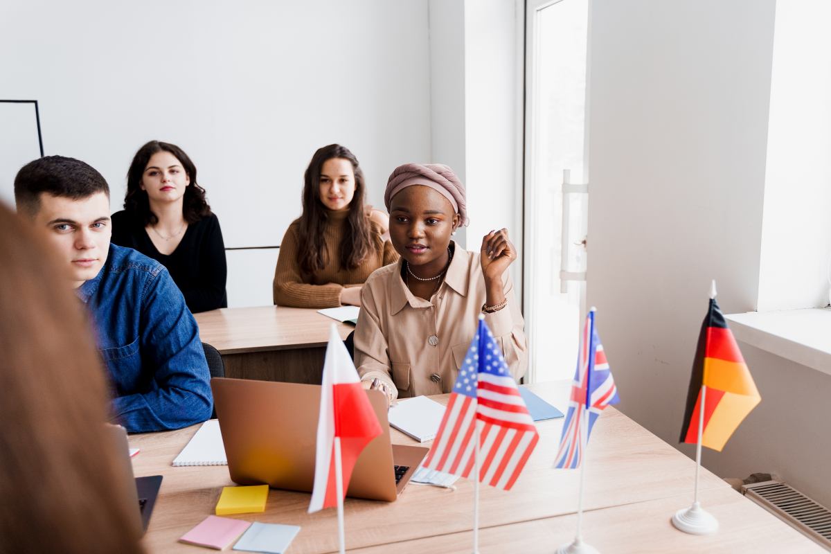 Gruppe internationaler Studierender sitzt mit Laptops und kleinen Länderflaggen an einem Tisch.