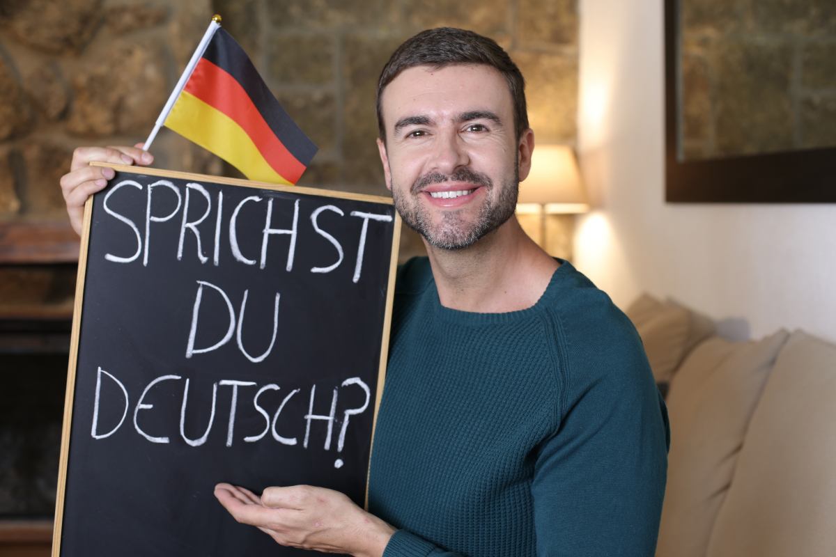 Mann hält eine kleine Deutschland-Flagge und eine Tafel mit der Aufschrift „Sprichst du Deutsch?“ in der Hand.