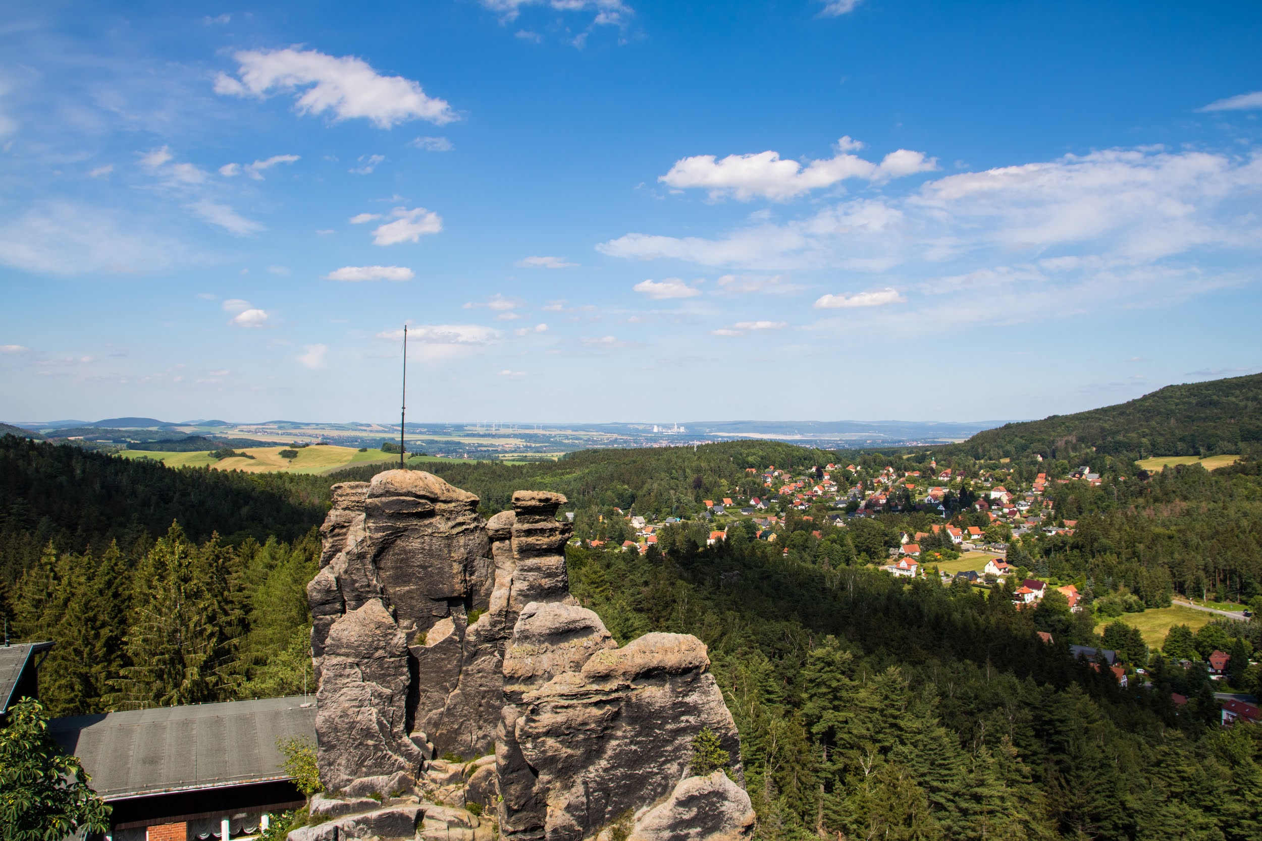 Weitblick auf das Zittauer Gebirge im Landkreis Görlitz. Im Vordergrund stehen große, graue Felsen. Eine dünne Antenne ragt von einem der Felsen in den Himmel. Hinter den Felsen erstreckt sich ein dichter grüner Wald. Dahinter liegt ein Dorf mit vielen roten Dächern und weißen Häusern, das sich über die Hügel zieht. Der Himmel ist blau mit einigen weißen Wolken.