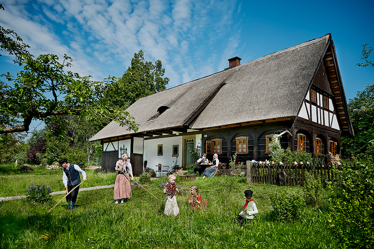 Zwei Erwachsene arbeiten in traditioneller Kleidung mit Sensen in einem Garten auf einer grünen Wiese. Drei Kinder in bunter Kleidung spielen daneben. Dahinter ist ein historisches Fachwerkhaus. Auf der Veranda sitzen ein Mann und eine Frau und unterhalten sich. Der Himmel ist blau mit weißen Wolken.