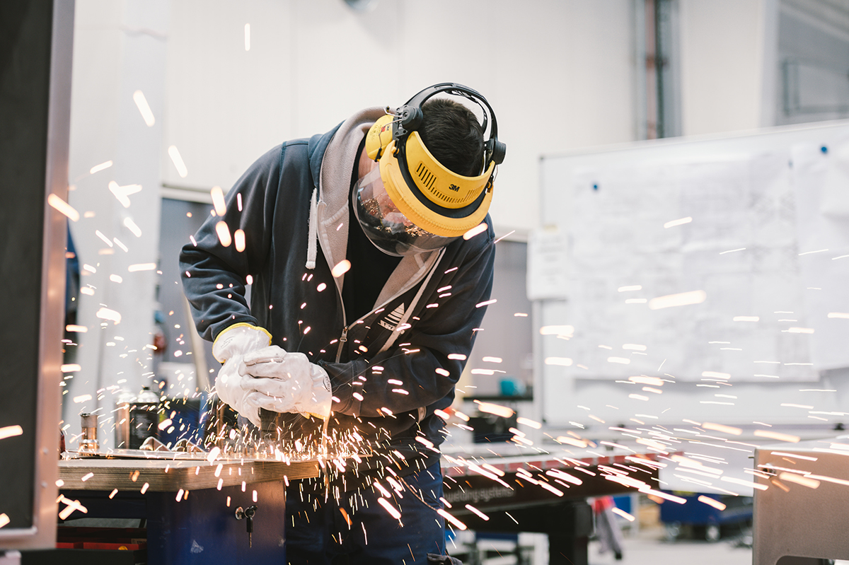 Eine Person arbeitet mit einer Schutzbrille und Gehörschutz an einem Werkstück. Funken fliegen in alle Richtungen. Die Person trägt eine dunkle Jacke und weiße Handschuhe. Der Arbeitsplatz ist eine Werkstatt mit Werkzeugen und Plänen im Hintergrund.