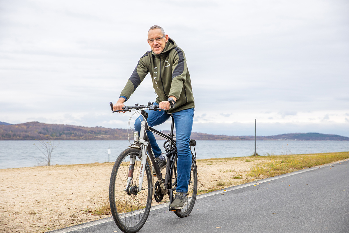 Auf dem Bild fährt ein Mann mit einem Fahrrad auf einem asphaltierten Weg am Ufer eines Sees entlang. Er trägt eine grüne Kapuzenjacke, Jeans und sportliche Schuhe. Der Mann lächelt und wirkt entspannt. Im Hintergrund sind ein Sandstrand, Wasser und bewaldete Hügel zu sehen. Der Himmel ist bewölkt, was auf einen kühlen Tag hindeutet. Das Bild vermittelt eine ruhige, naturnahe Stimmung und zeigt Freude an Bewegung im Freien.