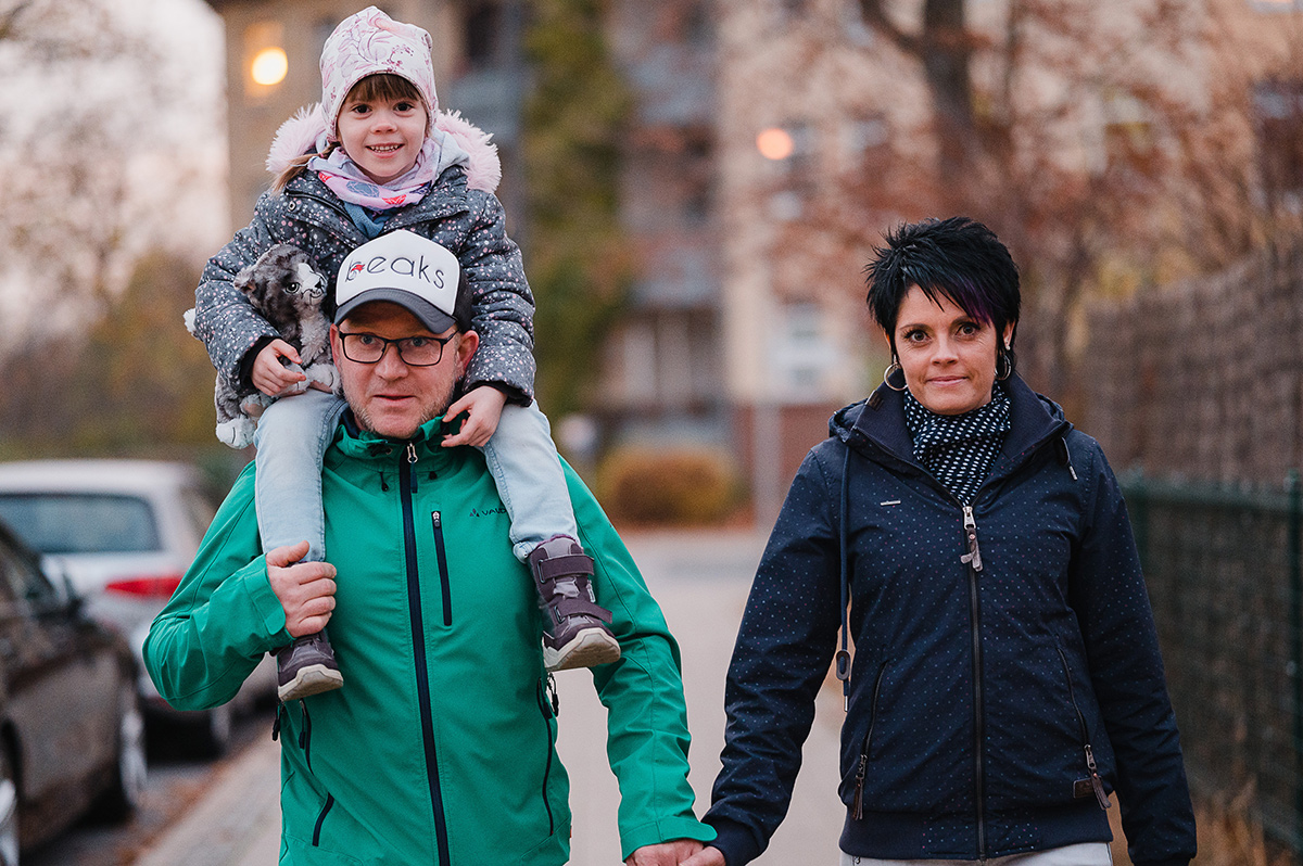Auf dem Bild ist eine Familie beim Spaziergang zu sehen. Ein Mann trägt ein kleines Mädchen auf seinen Schultern. Das Kind lächelt fröhlich, trägt eine Mütze, eine dicke Jacke und hält ein Kuscheltier im Arm. Der Mann trägt eine grüne Jacke, eine Brille und eine Mütze. Neben ihnen läuft eine Frau, die seine Hand hält. Sie trägt eine dunkelblaue Jacke, einen Schal und hat dunkles, kurzes Haar mit lila Strähnen. Im Hintergrund erkennt man eine herbstliche Umgebung mit kahlen Bäumen, geparkten Autos und Häusern. Das Bild vermittelt eine familiäre, gemütliche Stimmung.