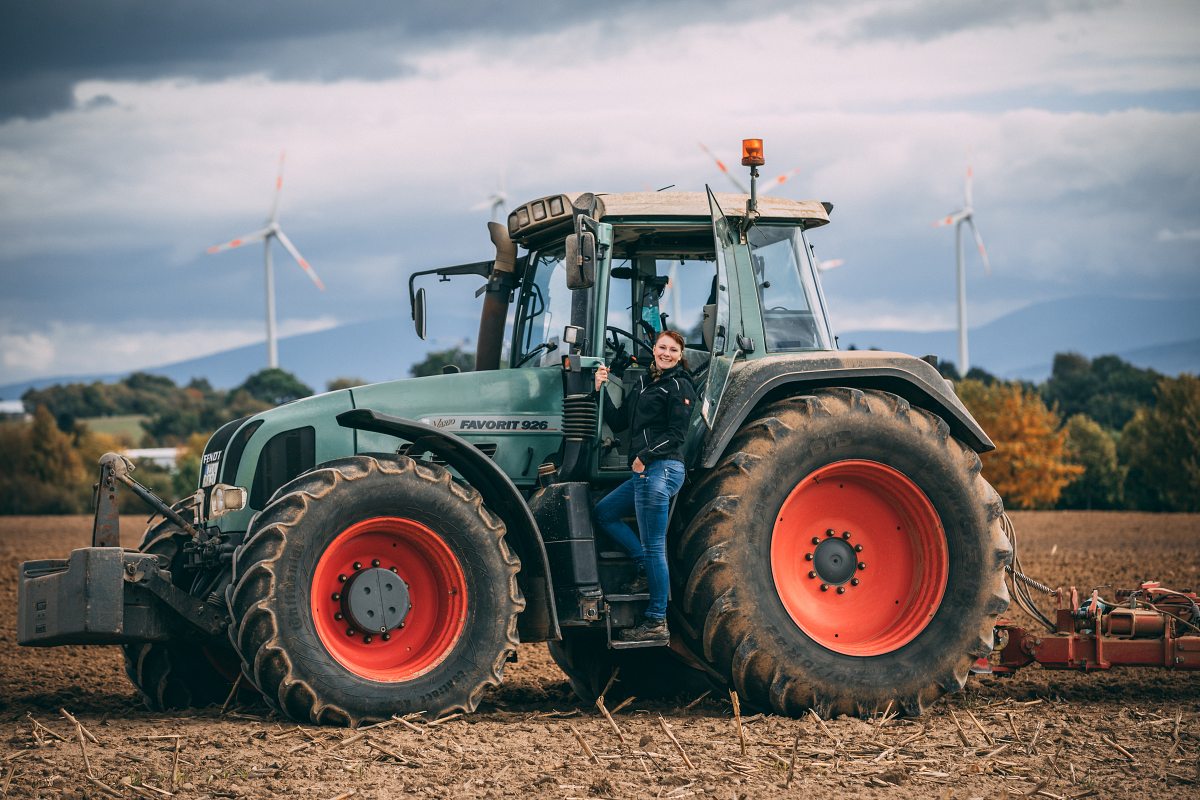 Eine Frau steht auf der Trittstufe eines großen grünen Traktors auf einem Feld. Sie lächelt in die Kamera. Im Hintergrund sind mehrere Windräder und Hügel zu sehen. Der Himmel ist bewölkt.