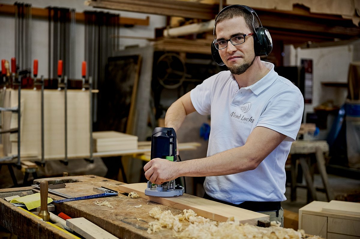Ein junger Mann mit Brille und Gehörschutz arbeitet in einer Werkstatt. Er bearbeitet mit einem technischen Gerät ein Stück Holz. Auf der Werkbank liegen Holzspäne und Werkzeuge. Im Hintergrund sind weitere Holzplatten und Werkzeuge zu sehen.
