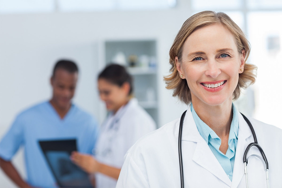 Eine lächelnde Ärztin steht im Vordergrund einer hellen Praxis oder Klinik. Im Hintergrund besprechen sich zwei medizinische Fachkräfte über ein Tablet. Das Bild vermittelt Professionalität, Vertrauen und Teamarbeit im Gesundheitswesen.