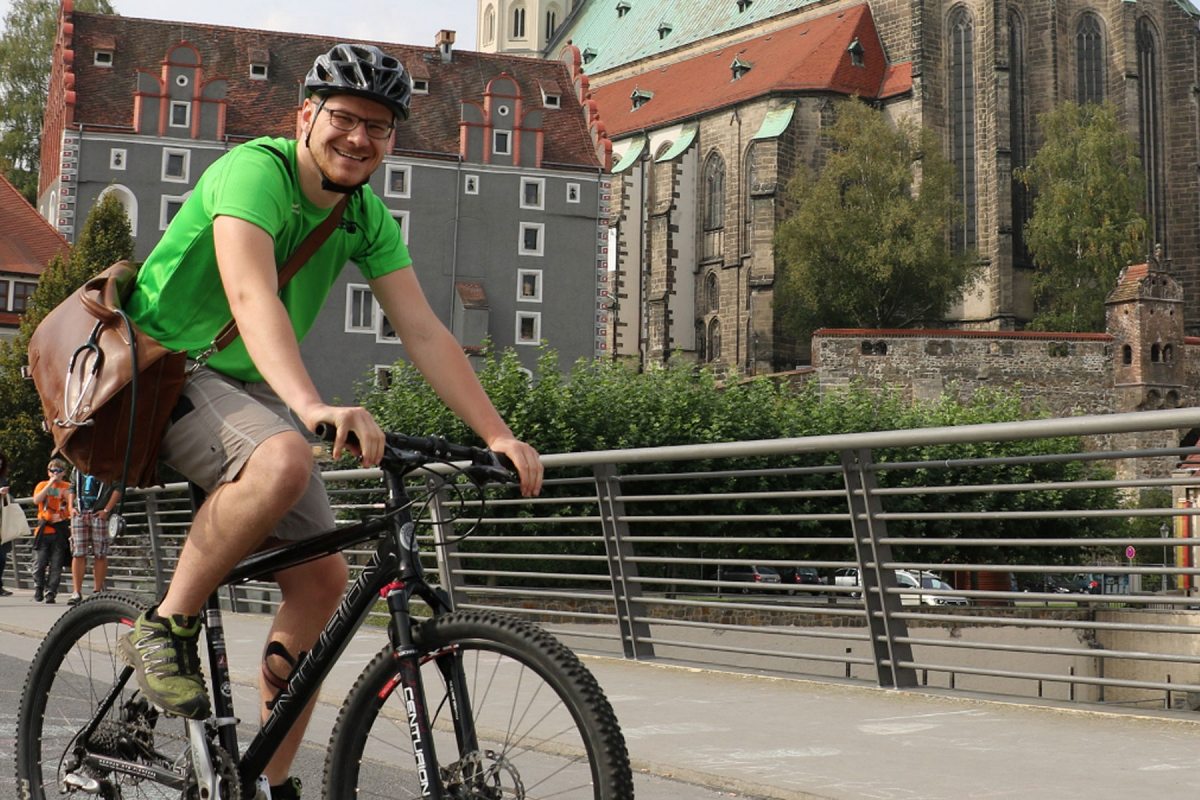 Ein Mann mit Helm und grünem T-Shirt fährt lächelnd auf einem Fahrrad über die Görlitzer Altstadtbrücke. Er trägt eine braune Umhängetasche. Im Hintergrund sind Teile einer Kirche und eines Gebäudes zu sehen.