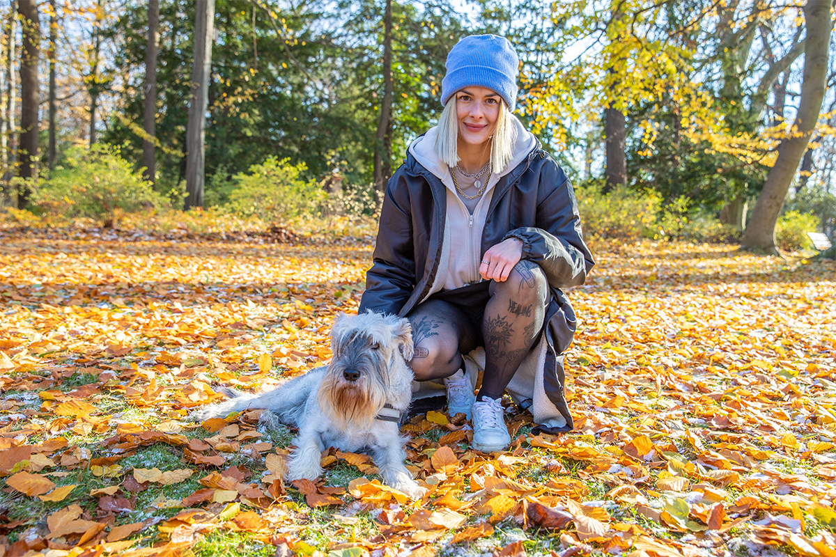 Eine junge Frau mit blonden Haaren, einer grauen Mütze, einer schwarzen Jacke und schwarzen durchsichtigen Strumpfhosen mit Tattoos hockt in einem Park auf der Wiese. Darauf liegen viele bunte Blätter. Neben ihr sitzt ein grauer, zotteliger Hund. Im Hintergrund sind grüne Bäume.