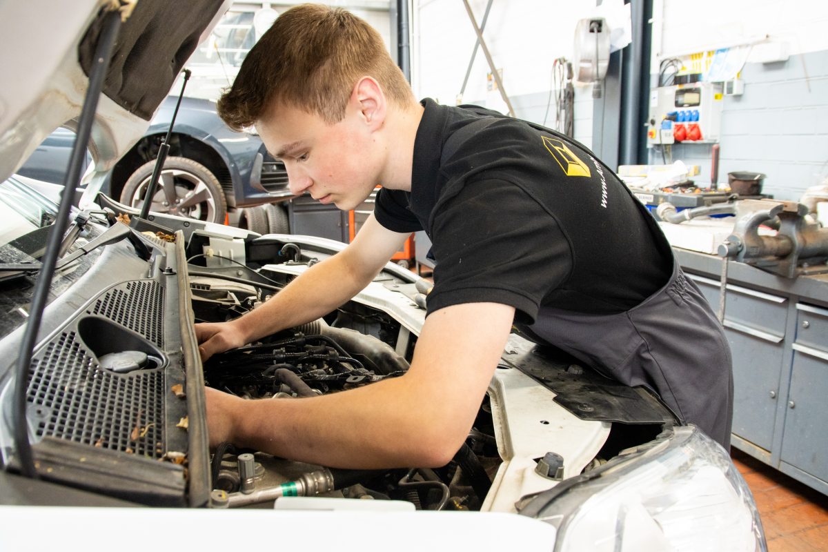 Ein junger Mann in Arbeitskleidung beugt sich über den offenen Motorraum eines Autos und repariert etwas. Er arbeitet in einer hellen Werkstatt mit Werkzeugen im Hintergrund.