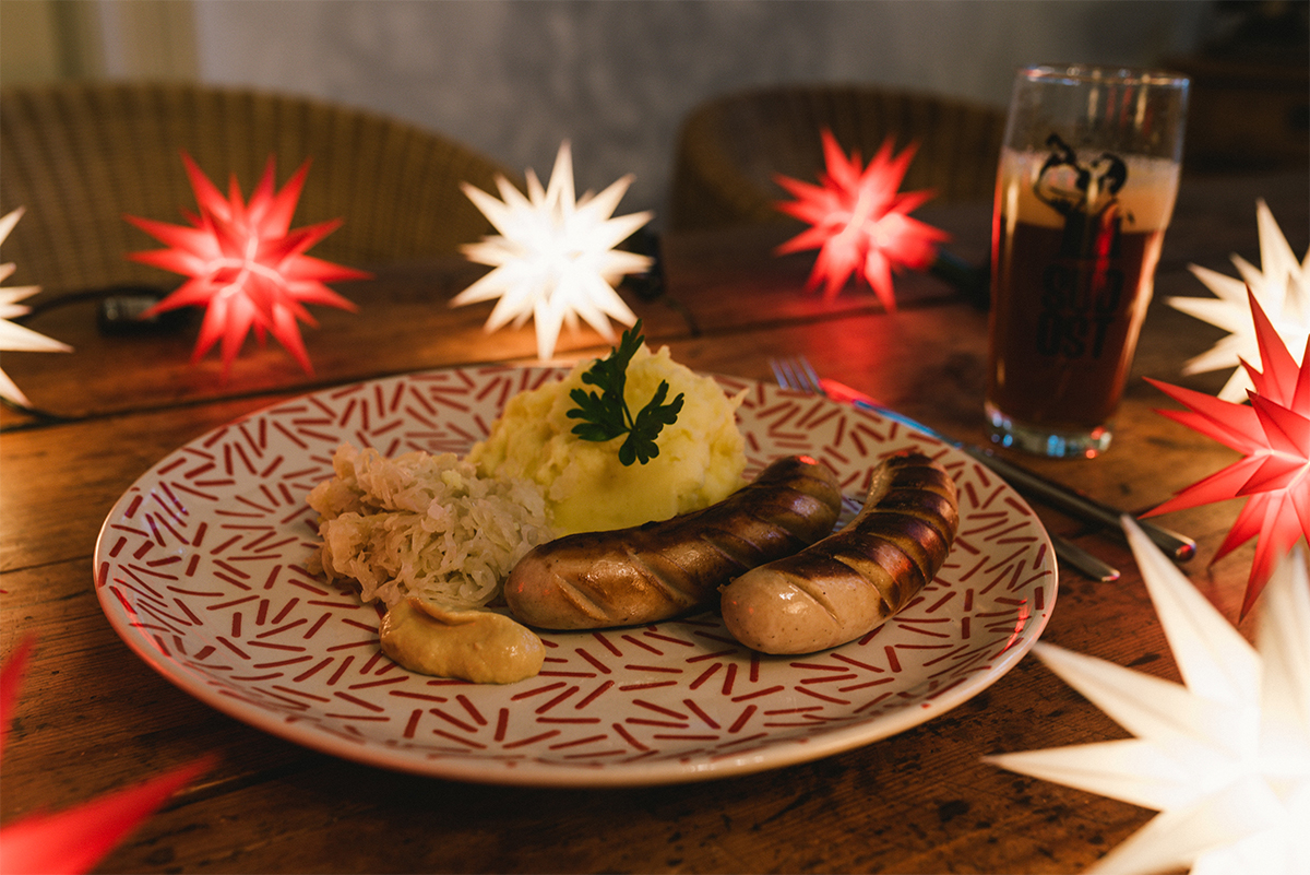 Das Bild zeigt ein traditionell angerichtetes deutsches Gericht auf einem dekorativen Teller mit rot-weißem Muster. Auf dem Teller liegen zwei gebratene Bratwürste mit deutlichen Grillstreifen, dazu eine Portion Sauerkraut und cremiges Kartoffelpüree, das mit einem Petersilienzweig garniert ist. Daneben befindet sich ein Klecks Senf. Der Tisch ist aus Holz und rustikal, im Hintergrund sorgen rot-weiße Weihnachtssterne (Herrnhuter Sterne) für eine gemütliche, festliche Beleuchtung. Rechts im Bild steht ein Glas mit dunklem Bier, auf dem ein Logo zu sehen ist. Der Gesamteindruck ist sehr stimmungsvoll und einladend, passend zu einem traditionellen, winterlichen Essen in gemütlicher Atmosphäre.