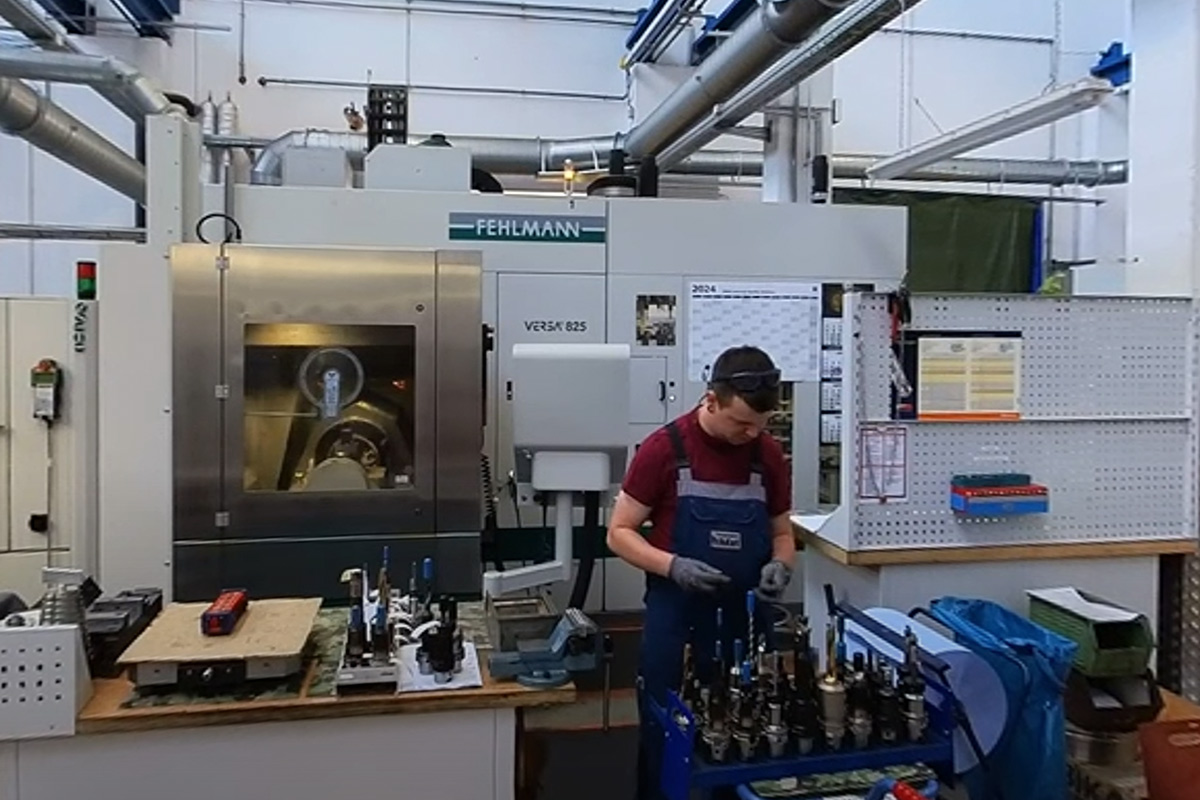 Ein junger Mann in blauer Latzhose steht vor einer großen Maschine mit der Aufschrift „FEHLMANN“ in einer Werkstatt. Er sortiert Metallwerkzeuge auf einem Wagen. Um ihn herum stehen Werkbänke, Maschinen und Arbeitsmaterialien.