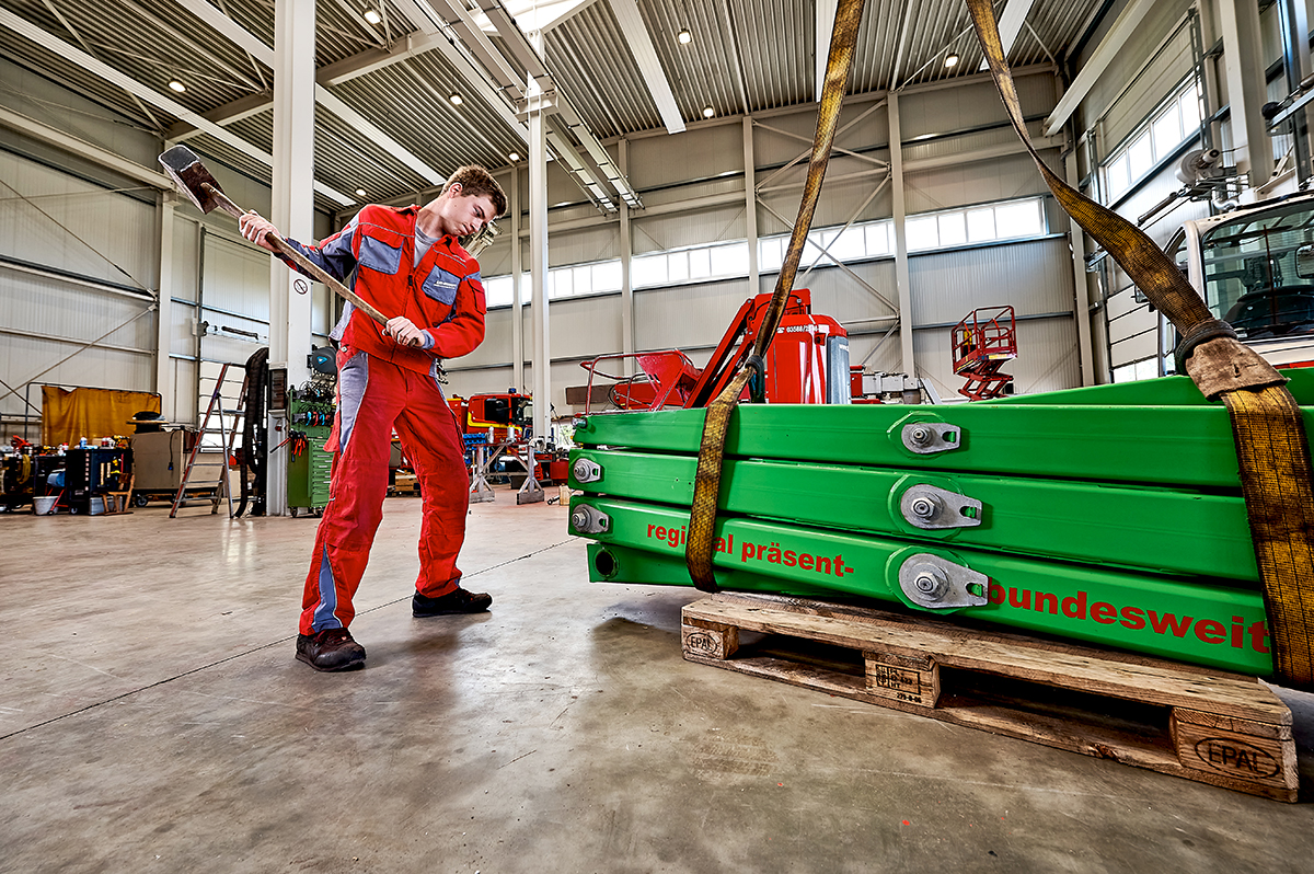 Auf dem Bild ist ein junger Mann in einer Werkshalle zu sehen, der in roter Arbeitskleidung kräftig mit einem großen Hammer auf ein grünes Metallteil schlägt. Das Metallteil liegt auf einer Palette und ist mit einem Spanngurt gesichert. Im Hintergrund sind weitere Maschinen, Werkzeuge und Arbeitsgeräte in der geräumigen, hellen Halle zu erkennen. Der Schriftzug auf dem Metallteil lautet „regional präsent – bundesweit“. Das Bild vermittelt handwerkliche Arbeit, körperlichen Einsatz und eine produktive Werkstatt-Atmosphäre.