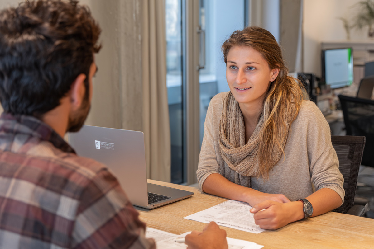 Zwei Personen sitzen sich an einem Schreibtisch gegenüber. Die junge Frau hört aufmerksam zu, während der Mann Unterlagen in der Hand hält. Ein Laptop steht zwischen ihnen. Die Szene wirkt wie ein Beratungsgespräch in einer professionellen Umgebung.