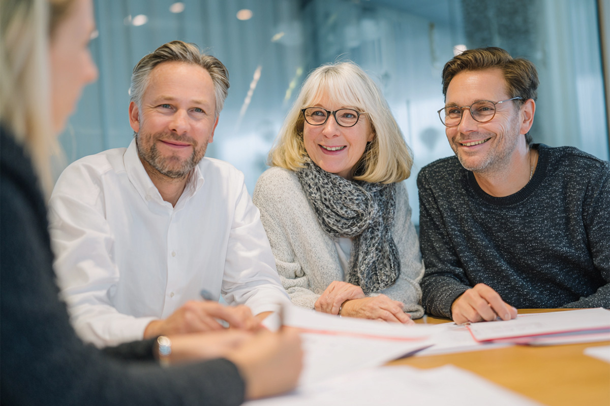 Drei Personen sitzen lächelnd an einem Tisch mit Unterlagen und schauen eine vierte Person an. Die Szene wirkt freundlich und professionell – sie symbolisiert ein Beratungsgespräch, z. B. im Rahmen einer Gründungsberatung bei der IHK oder einer ähnlichen Institution.