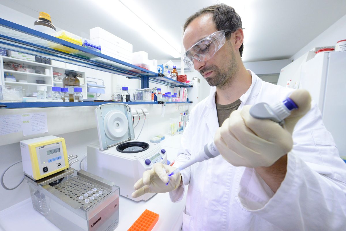 Ein Mann in weißem Laborkittel und Schutzbrille arbeitet mit einer Pipette in einem Labor. Um ihn herum stehen Laborgeräte. Der Raum ist hell und modern ausgestattet.