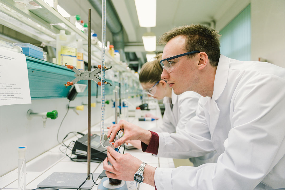 Ein junger Mann und eine junge Frau in weißen Laborkitteln und Schutzbrillen arbeiten konzentriert in einem Chemielabor. Sie führen Messungen mit Glasinstrumenten durch. Im Hintergrund stehen viele Laborgeräte und Flaschen mit Chemikalien.