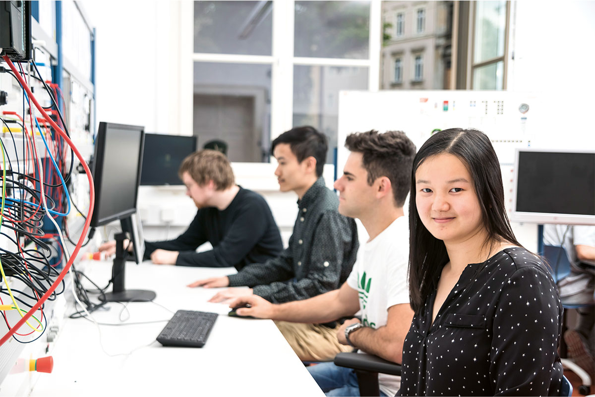 Mehrere junge Erwachsene arbeiten an Computern in einem modernen Technikraum. Links sind viele bunte Kabel an einer Wand angebracht. Eine junge Frau im Vordergrund lächelt in die Kamera. Im Hintergrund schauen die anderen konzentriert auf ihre Bildschirme.