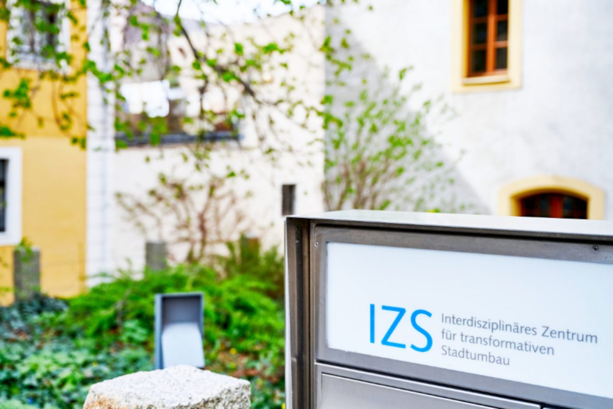 Ein Schild vor einem Gebäude zeigt den Schriftzug „IZS – Interdisziplinäres Zentrum für transformativen Stadtumbau“. Im Hintergrund sind grüne Pflanzen und Hausfassaden mit Fenstern zu sehen.
