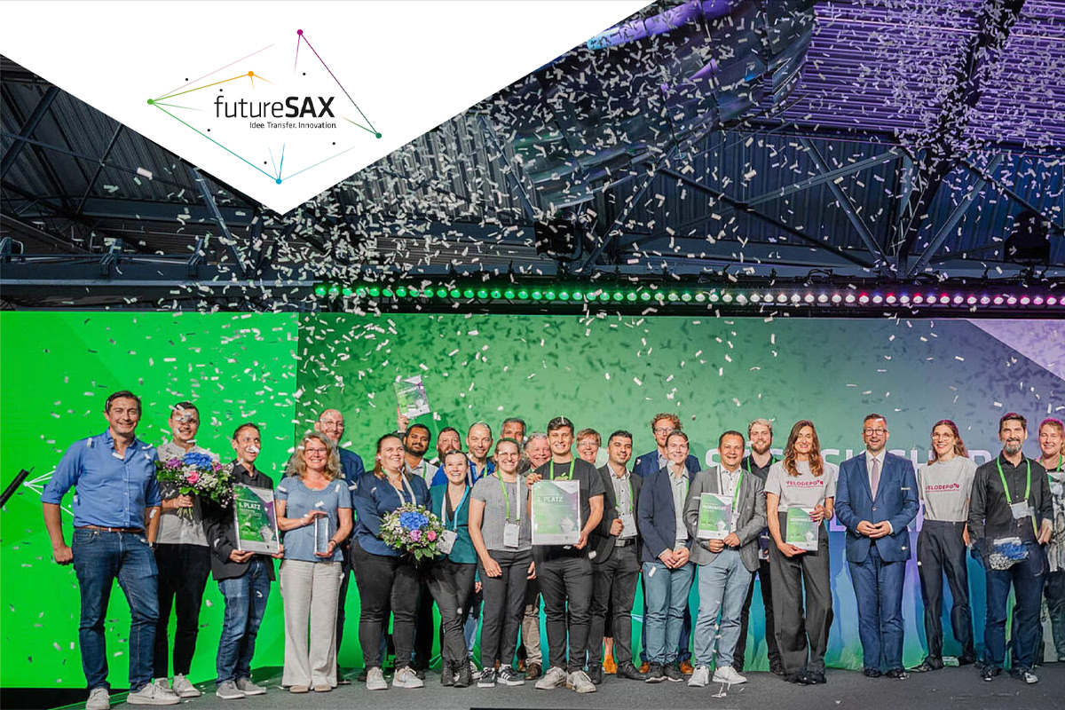 Gruppenbild von rund 25 Personen auf einer Bühne bei einer Preisverleihung von futureSAX – dem Innovationsnetzwerk des Freistaates Sachsen. Viele lächeln, halten Urkunden oder Blumensträuße in der Hand. Über ihnen fällt Konfetti herab. Im Hintergrund ist ein leuchtend grüner Bühnenhintergrund mit farbiger Lichtleiste. Oben links ist das futureSAX-Logo mit dem Slogan „Idee. Transfer. Innovation.“ eingeblendet.