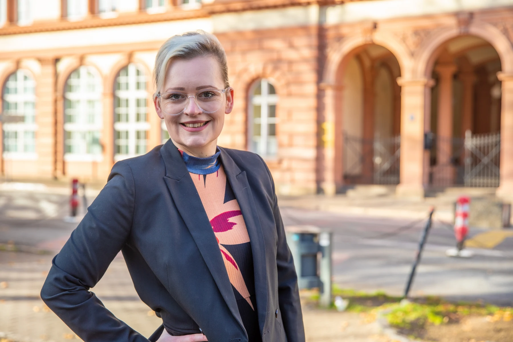 Auf dem Bild steht eine Frau im Freien vor einem historischen Gebäude mit Rundbögen und großen Fenstern. Sie trägt eine auffällige Brille, ein gemustertes Oberteil und einen dunklen Blazer. Mit einer Hand in der Hüfte und einem offenen, selbstbewussten Lächeln blickt sie direkt in die Kamera. Die warme Beleuchtung und die Architektur im Hintergrund vermitteln eine freundliche, professionelle Atmosphäre – möglicherweise im Kontext von Bildung oder öffentlicher Verwaltung.