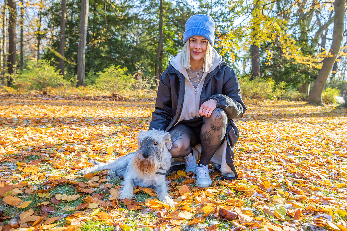 Auf dem Bild sitzt eine junge Frau an einem herbstlich bedeckten Waldboden, umgeben von buntem Laub. Sie trägt eine blaue Mütze, eine helle Kapuzenjacke, eine dunkle Oversize-Jacke und weiße Sneaker. Ihre tätowierten Beine sind sichtbar, und sie lächelt freundlich in die Kamera. Neben ihr sitzt ein kleiner, zotteliger Hund mit hellem Fell, der ebenfalls aufmerksam in Richtung Kamera blickt. Die Stimmung ist entspannt und lebendig – ein Moment der Verbundenheit mit der Natur und dem Haustier an einem sonnigen Herbsttag.