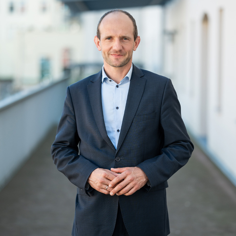 Dr. Stephan Meyer, Landrat des Landkreises Görlitz, steht in einem hellen Hemd und einem dunklen Anzug auf einem Balkon. Er lächelt leicht und hat die Hände vor dem Körper gefaltet.