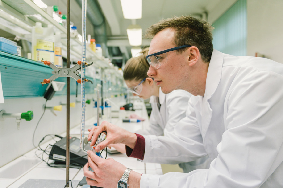 Ein junger Mann und eine junge Frau in weißen Laborkitteln und Schutzbrillen arbeiten konzentriert in einem Chemielabor. Sie führen Messungen mit Glasinstrumenten durch. Im Hintergrund stehen viele Laborgeräte und Flaschen mit Chemikalien.