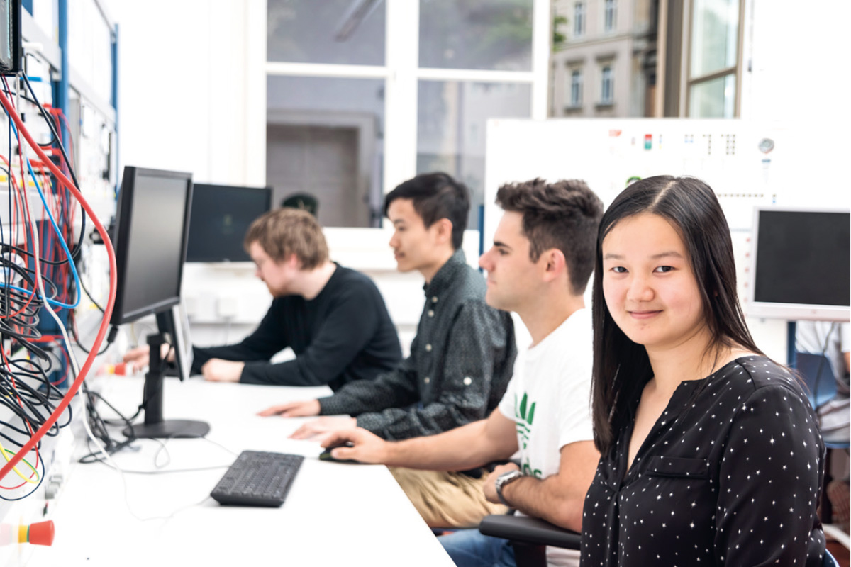 Mehrere junge Erwachsene arbeiten an Computern in einem modernen Technikraum. Links sind viele bunte Kabel an einer Wand angebracht. Eine junge Frau im Vordergrund lächelt in die Kamera. Im Hintergrund schauen die anderen konzentriert auf ihre Bildschirme.