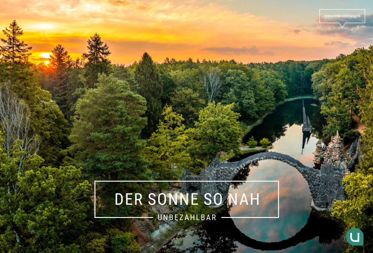 Die Rakotzbrücke in Kromlau. Auf dem Bild ist eine bewaldete Flusslandschaft bei Sonnenuntergang zu sehen. Eine halbkreisförmige Steinbrücke überspannt im rechten unteren Bereich einen ruhigen Fluss, in dem sich sowohl die Brücke als auch der orange-gelb gefärbte Himmel spiegeln. Der Fluss ist von dichtem, grünem Mischwald umgeben. Der Himmel zeigt leichte Bewölkung und ist von warmem Abendlicht gefärbt. Im unteren Bereich des Bildes befindet sich ein weiß umrahmter Text, in dem steht: „Der Sonne so nah“. Direkt darunter steht in kleinerer Schrift das Wort „Unbezahlbar“. In der oberen rechten Bildecke ist in einem weißen Rahmen die Webadresse „www.unbezahlbar.land“ zu lesen. Unten rechts ist ein rundes Logo mit einem weißen „u“ auf grünem Hintergrund abgebildet.