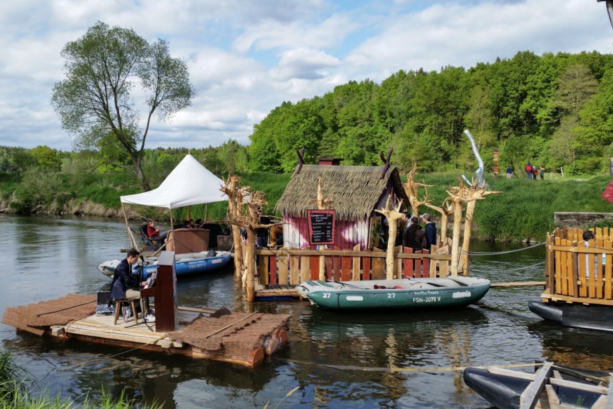 Mehrere Flöße und Boote befinden sich auf einem Fluss. Auf einem kleinen Floß sitzt ein Mann an einem Klavier. Ein anderes Floß trägt ein hölzernes Häuschen mit Reetdach und bunter Bemalung. An der Vorderseite des Hauses hängt eine schwarze Tafel mit Speisen und Getränken. Um das Floß stehen kunstvoll gestaltete Baumskulpturen. Im Hintergrund sind Bäume, Wiese und ein paar Menschen auf einem Pfad zu sehen. Der Himmel ist teils bewölkt.