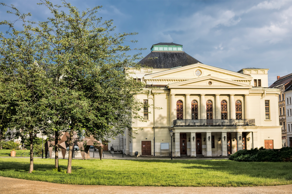 Das Gerhart Hauptmann- Theater in Görlitz. Auf dem Bild ist ein großes Gebäude mit Säulen und einer hellgelben Fassade zu sehen. Vor dem Gebäude stehen mehrere Bäume und ein Kunstobjekt aus Metall. Der Bereich vor dem Haus ist von einem Weg und einer gepflegten Rasenfläche umgeben. Im Hintergrund sind weitere Häuser zu erkennen. Der Himmel ist teilweise bewölkt.