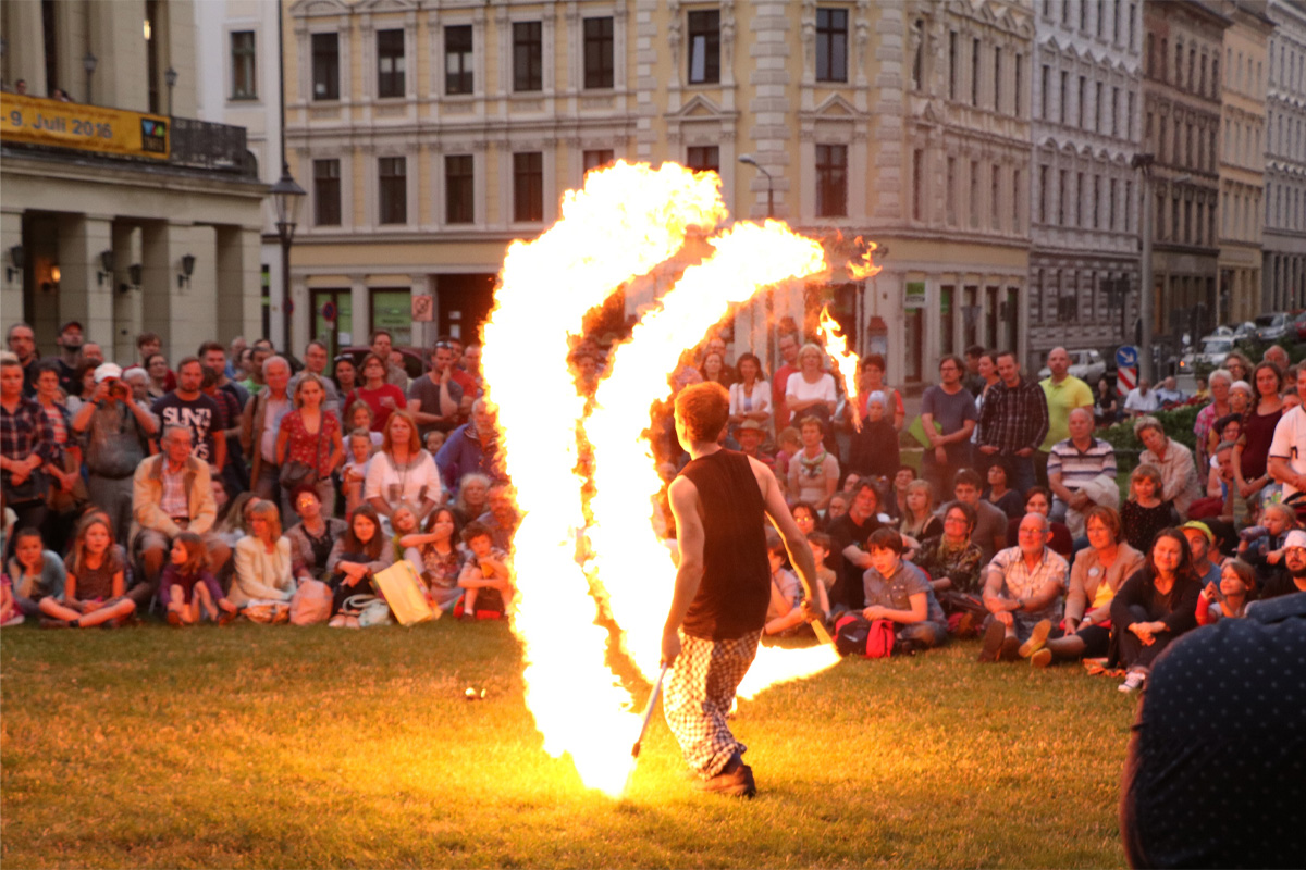 Auf dem Bild ist eine Feuershow im Freien zu sehen. Eine Person steht auf einer Wiese und hält zwei brennende Fackeln in der Hand. Um sie herum bilden die Flammen zwei deutliche Bögen. Im Hintergrund sitzt und steht eine große Menschenmenge dicht gedrängt und schaut zu. Im Hintergrund sind Gebäude zu sehen und es ist dämmrig.