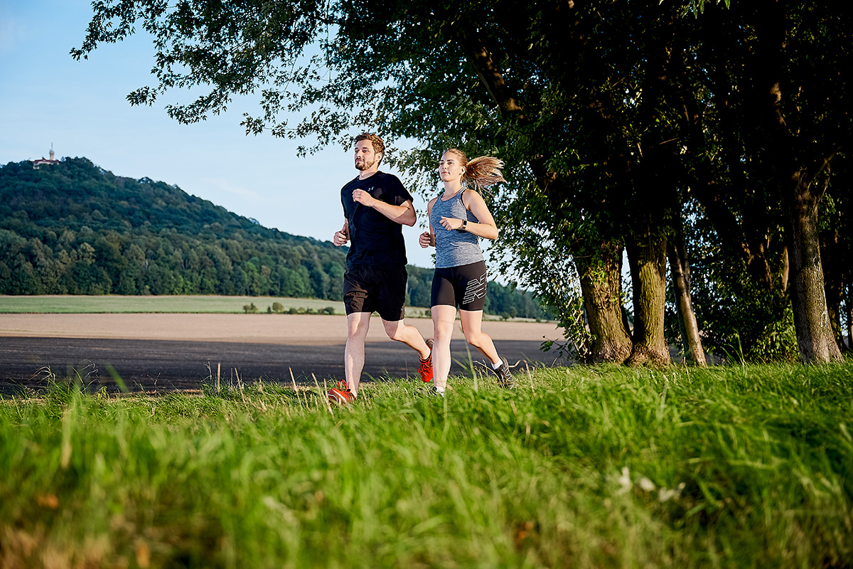 Ein Mann und eine Frau joggen nebeneinander auf einem grasbewachsenen Weg. Beide tragen sportliche Kleidung, der Mann in Schwarz und die Frau in Grau und Schwarz. Im Hintergrund ist eine weite Ackerlandschaft mit einem bewaldeten Hügel, der Landeskrone, zu sehen. Auf dem Hügel steht ein Gebäude mit einem Turm. Rechts im Bild befinden sich mehrere Bäume. Der Himmel ist klar.