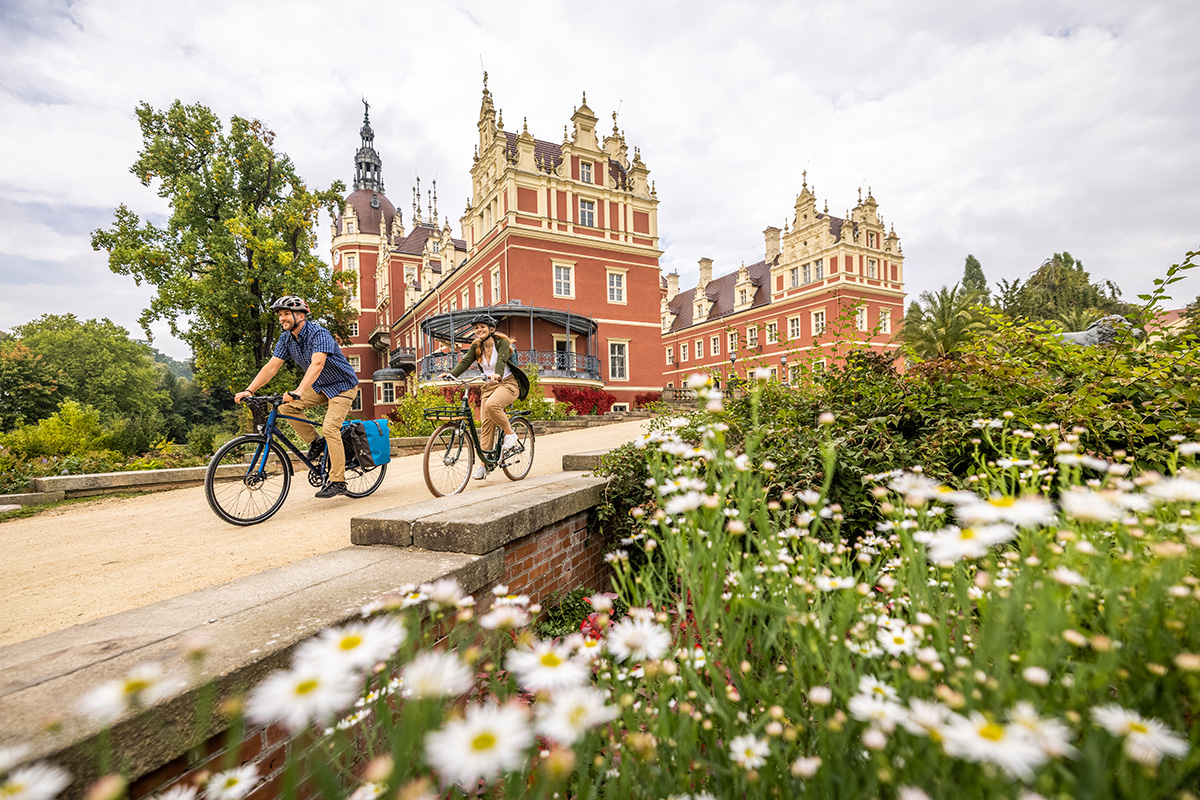 Das Schloss Muskau. Zwei Personen fahren auf Fahrrädern einen breiten Weg entlang, der an einem roten historischen Gebäude mit reich verzierten Türmen und Fassaden vorbeiführt. Im Vordergrund sind weiße Blüten einer Blumenwiese zu sehen. Der Weg verläuft auf einer niedrigen Brücke aus Stein, die von einem Ziegelmauerwerk gestützt wird. Im Hintergrund stehen große Bäume mit grünem Laub.