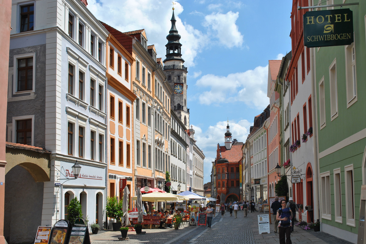 Die Brüderstraße in Görlitz. Zu sehen ist eine gepflasterte Straße in einer historischen Altstadt mit bunt gestalteten Fassaden auf beiden Seiten. Mehrere Tische mit Sonnenschirmen sind vor Restaurants aufgebaut, an denen Menschen sitzen. Im Hintergrund sind zwei hohe Türme mit spitzem Dach zu sehen. An einer Hauswand rechts hängt ein Schild mit der Aufschrift „Hotel Schwibbogen“. Es sind zahlreiche Passanten unterwegs. Der Himmel ist blau mit einigen Wolken.