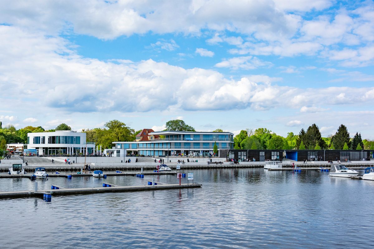 Es ist eine moderne Uferpromenade mit mehreren hellen Gebäuden mit großen Glasfronten zu sehen. Im Vordergrund befinden sich Stege und Bootsanleger mit kleinen Booten auf dem Wasser. Entlang der Promenade sind zahlreiche Personen zu sehen. Im Hintergrund sind Bäume zu erkennen, darüber ein teilweise bewölkter Himmel.