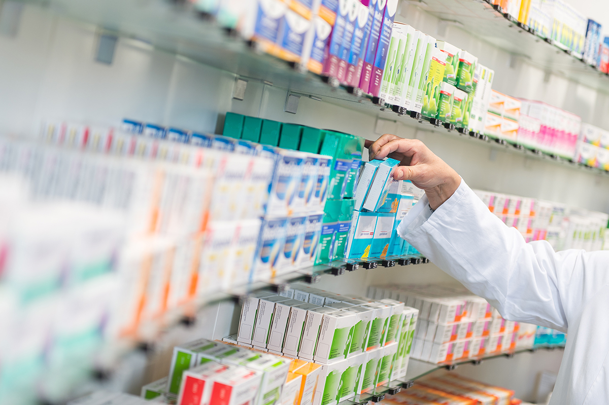 Ein Regal in einer Apotheke ist gefüllt mit bunten Medikamentenverpackungen, die sorgfältig nach Sorten geordnet sind. Im rechten Bildbereich greift ein Arm in weißem Kittel – vermutlich von einem Apotheker oder einer Apothekerin – nach einer türkisfarbenen Arzneimittelpackung in der mittleren Reihe.