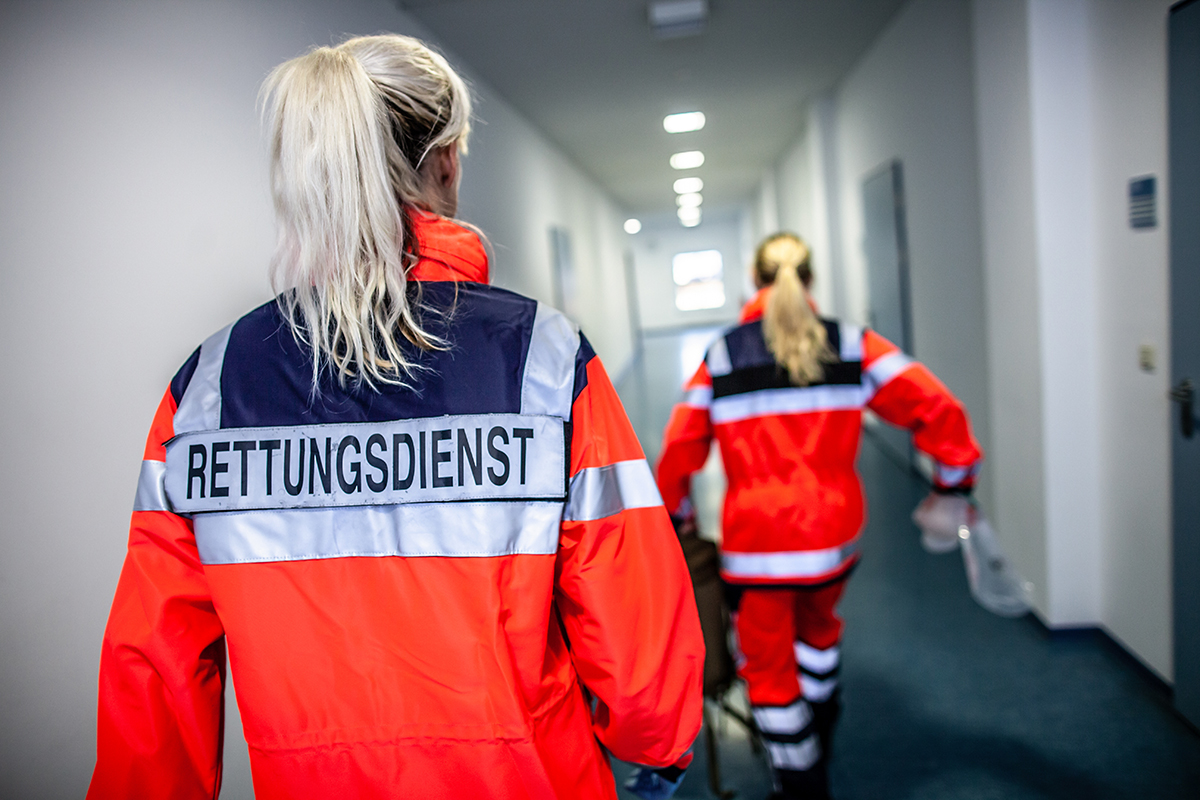 Zwei Frauen in leuchtend orange-roter Einsatzkleidung mit der Aufschrift „RETTUNGSDIENST“ gehen durch einen hellen Flur. Die hintere Person ist unscharf und trägt ein medizinisches Gerät in der Hand. Beide haben blonde, zum Zopf gebundene Haare. Die Szene ist aus der Rückansicht aufgenommen.