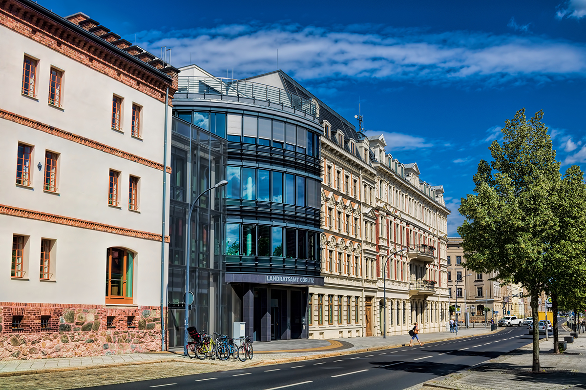 Auf dem Bild ist eine Straßenansicht mit zwei Gebäuden zu sehen. Links befindet sich ein modernes Gebäude mit abgerundeter Glasfassade und dunkler Verkleidung, darüber ist in großen Buchstaben „LANDRATSAMT GÖRLITZ“ zu lesen. Daneben steht ein historisches Gebäude mit reich verzierter Fassade in Beige und Rot. Im Vordergrund verläuft eine breite Straße mit Gehweg, am rechten Bildrand ragen grüne Bäume ins Bild. Der Himmel ist teilweise von Wolken bedeckt.